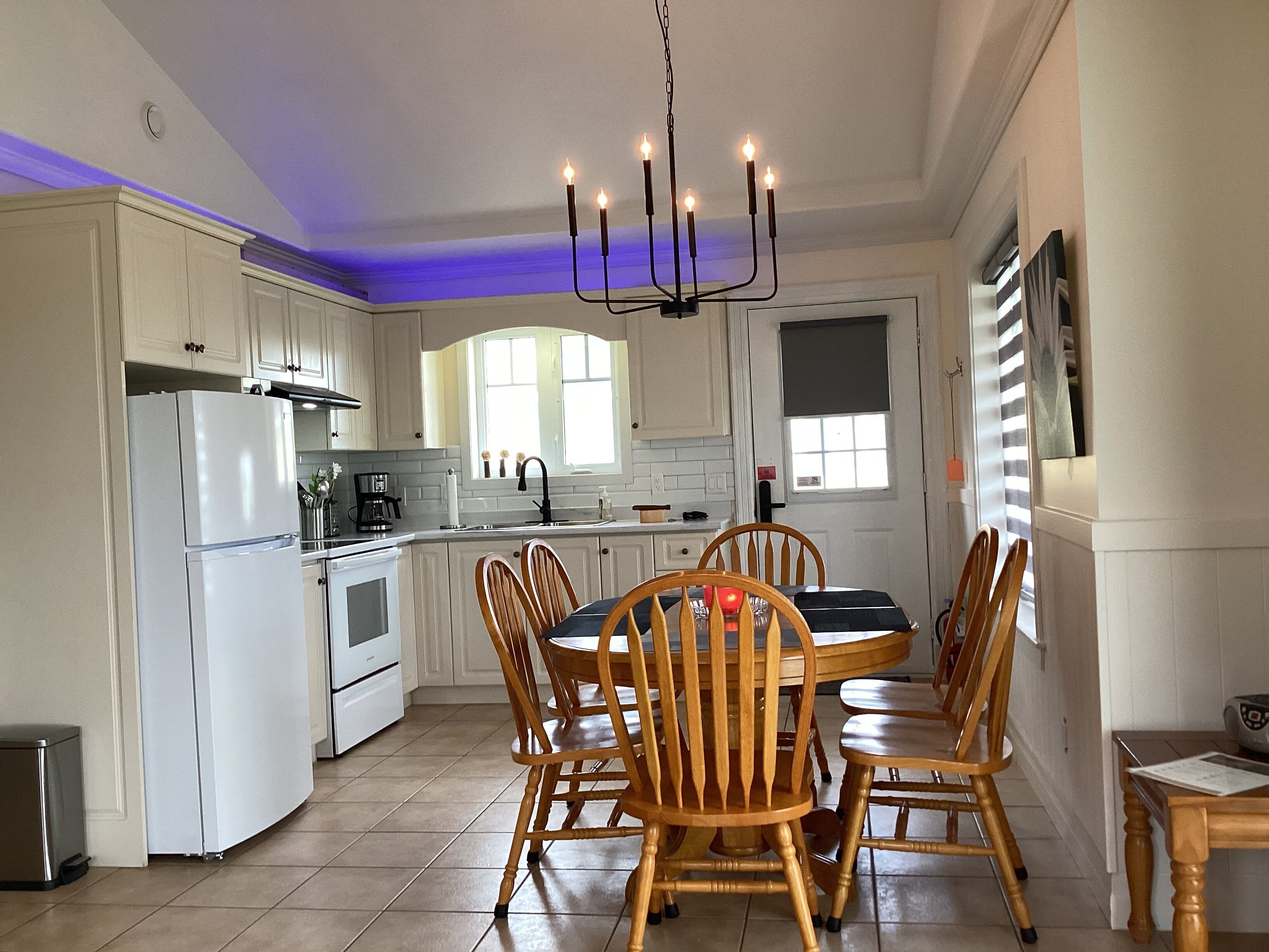 Una cocina con armarios blancos, un refrigerador blanco, una mesa de comedor con sillas de madera y un candelabro.