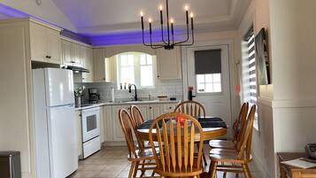 Una cocina con armarios blancos, un refrigerador blanco, una mesa de comedor con sillas de madera y un candelabro.
