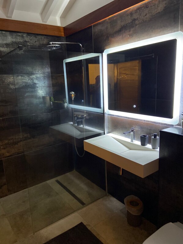 A modern bathroom with a glass shower enclosure, a white sink with a mirror, and a dark tiled wall.