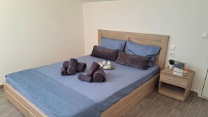 Ein ordentlich bezogenes Bett mit blauer Bettdecke, zwei Kissen und gerollten Handtüchern. Ein Nachttisch mit einer kleinen Pflanze und einem gerahmten Bild sind ebenfalls zu sehen.