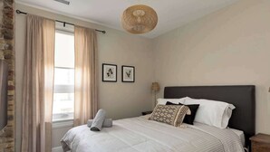 Een slaapkamer met een groot bed, beige gordijnen, een geweven plafondlamp en lijstwerken aan de wand.