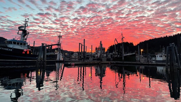 Un porto al tramonto con una barca chiamata "ANTHEM" ormeggiata, acqua calma che riflette il cielo, e un molo con diverse strutture.