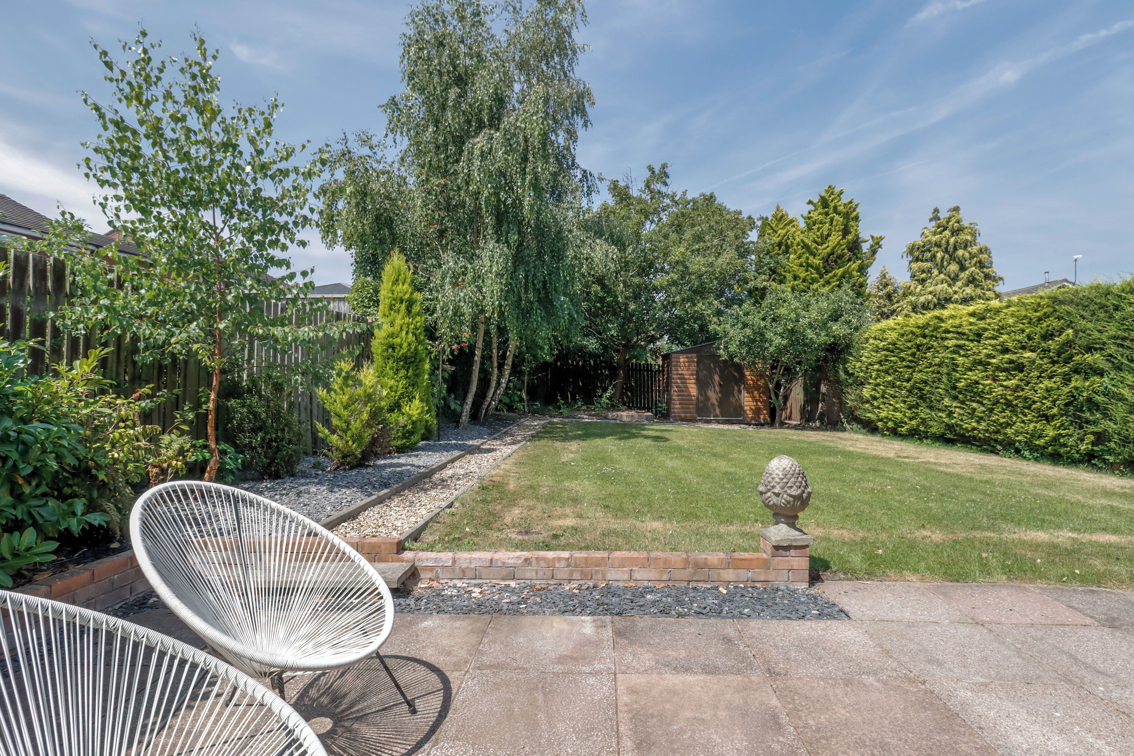 Un giardino curato con un'area patio, una sedia bianca e un sentiero in pietra che conduce a una struttura in legno.