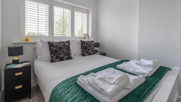 A neatly made bed with white linens, a green throw blanket, and white towels. There are two bedside tables with lamps and a small nightstand with a lamp. The room has white walls and a window with blinds. - Stylish Cheshire House (Winsford)