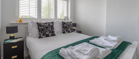 A neatly made bed with white linens, a green throw blanket, and white towels. There are two bedside tables with lamps and a small nightstand with a lamp. The room has white walls and a window with blinds.