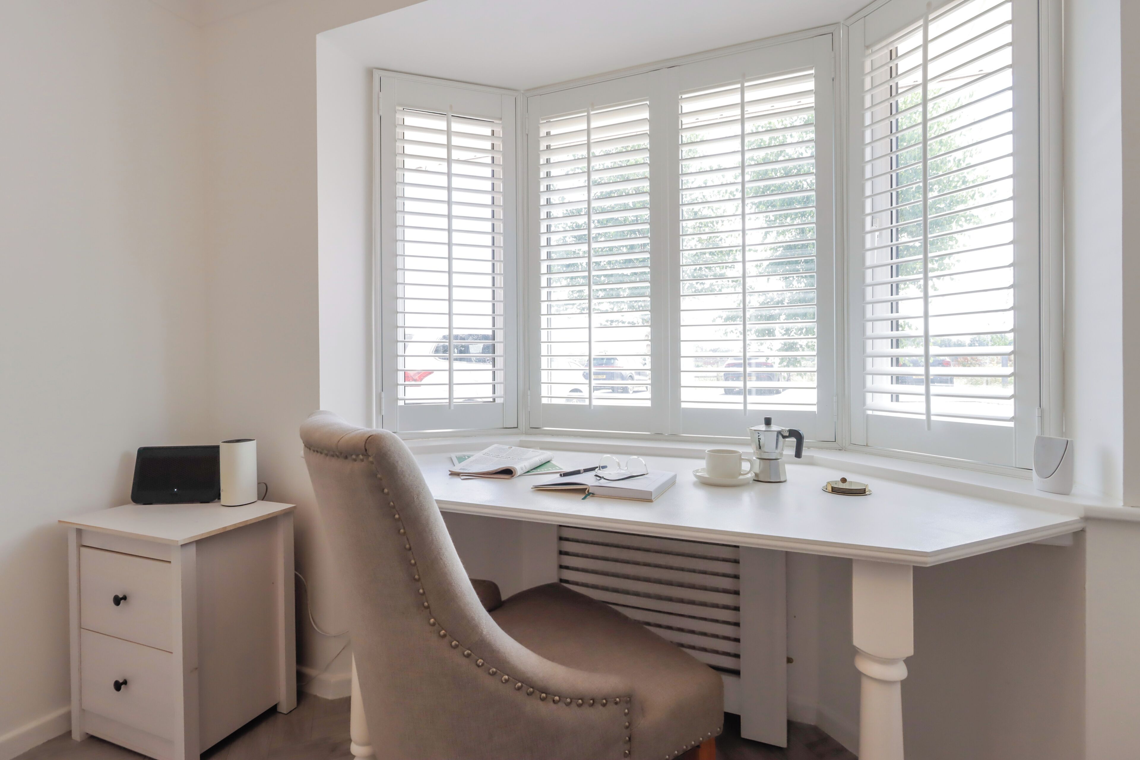 Un bureau d’angle avec une chaise, un petit meuble à tiroirs et une fenêtre avec des stores.