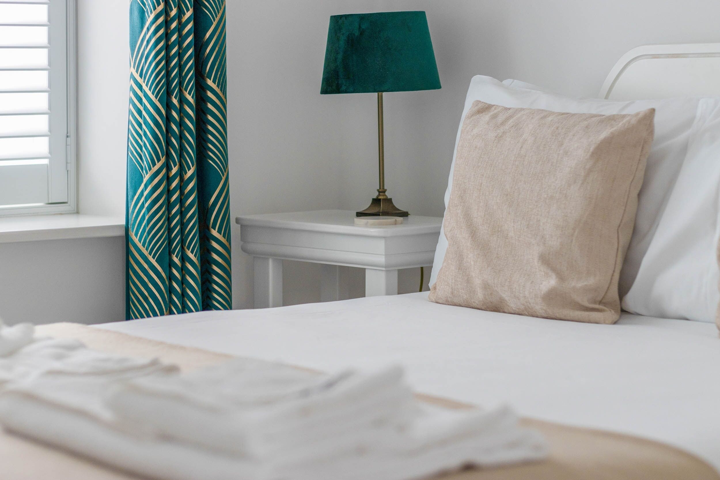 A neatly made bed with a white pillow and a beige pillow, a green lamp on a bedside table, and a window with blinds.