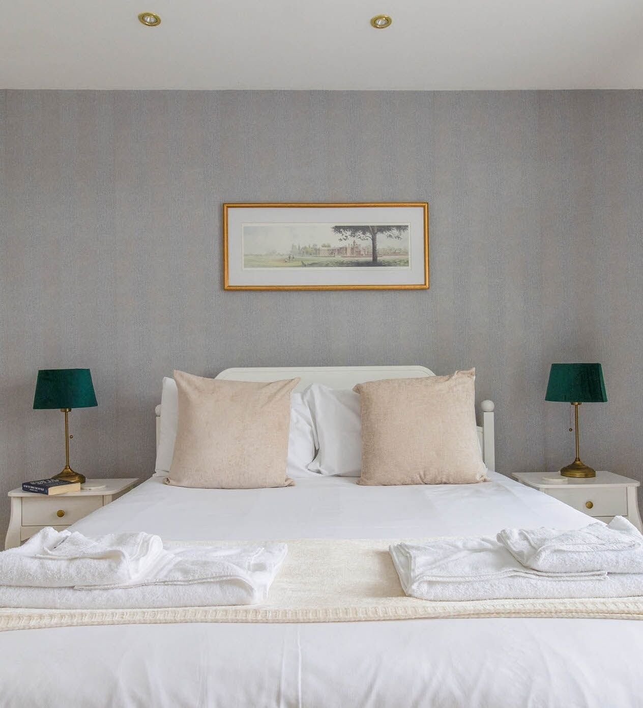 A neatly made bed with white linens and two pillows, flanked by two bedside tables with lamps, in a room with a grey textured wall and a framed picture above the bed.
