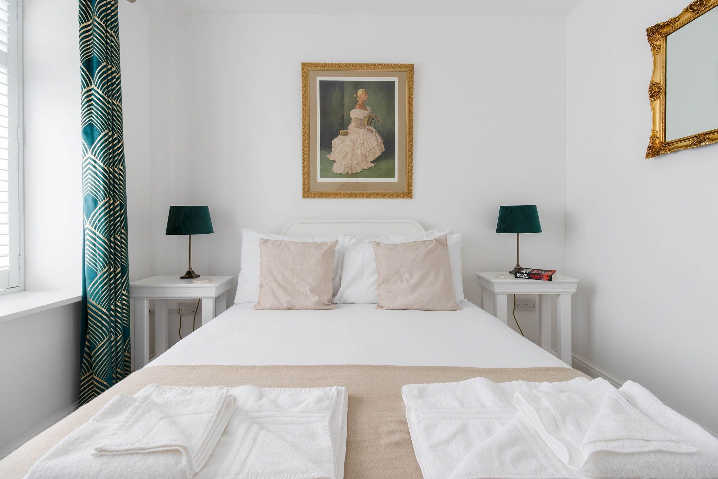 A neatly made bed with white linens, two bedside tables with lamps, a framed painting on the wall, and a window with curtains.