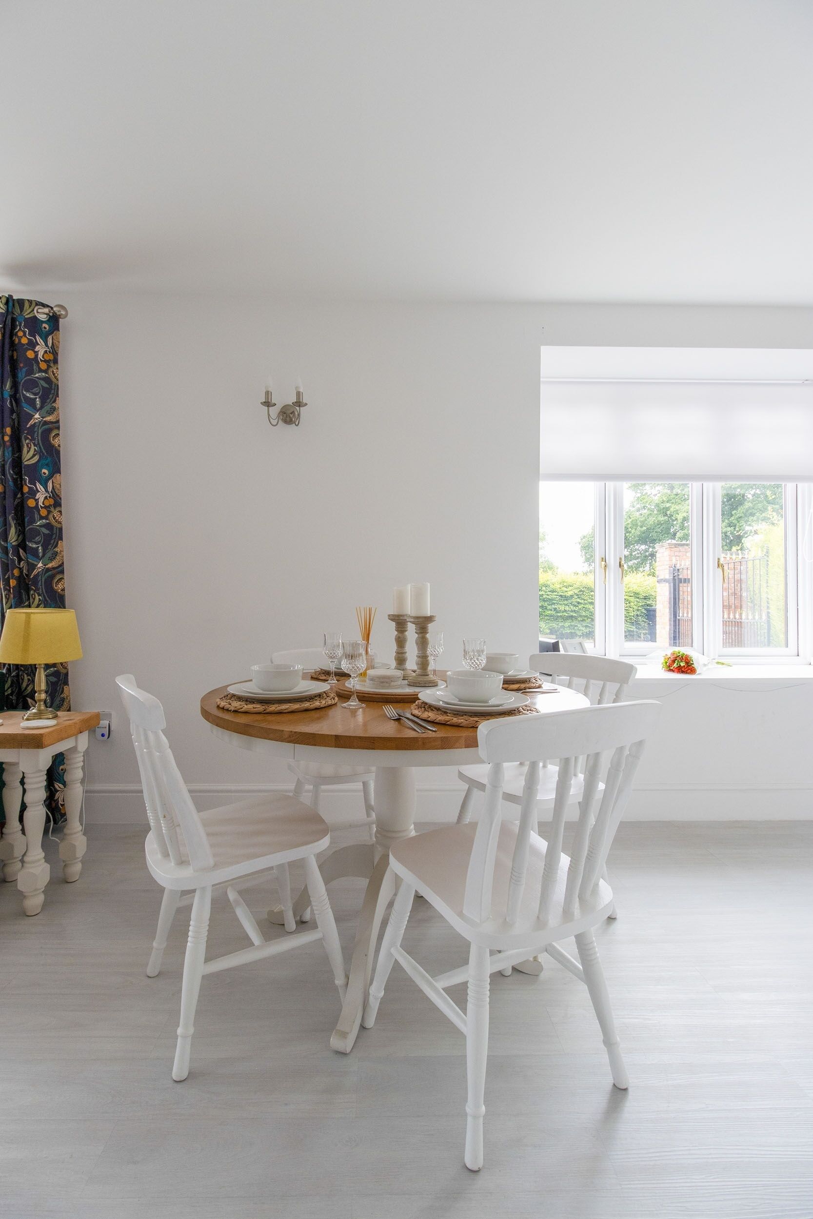 Een eetruimte met een ronde tafel gedekt voor vier personen, witte stoelen, een lamp en een raam met uitzicht op groen.