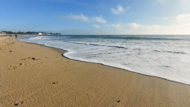 A sandy beach with gentle waves, a clear sky, and a distant shoreline with buildings.