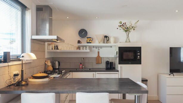 Una cucina moderna con piano di lavoro scuro, mobili bianchi, forno a microonde integrato e un piccolo televisore.