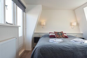 Een slaapkamer met een schuine kap, twee bedlampen, een bed met een grijze dekbedovertrek, en een raam met zonwering.