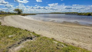 Uma praia arenosa com um corpo d'água calmo, cercada por áreas gramadas e árvores.