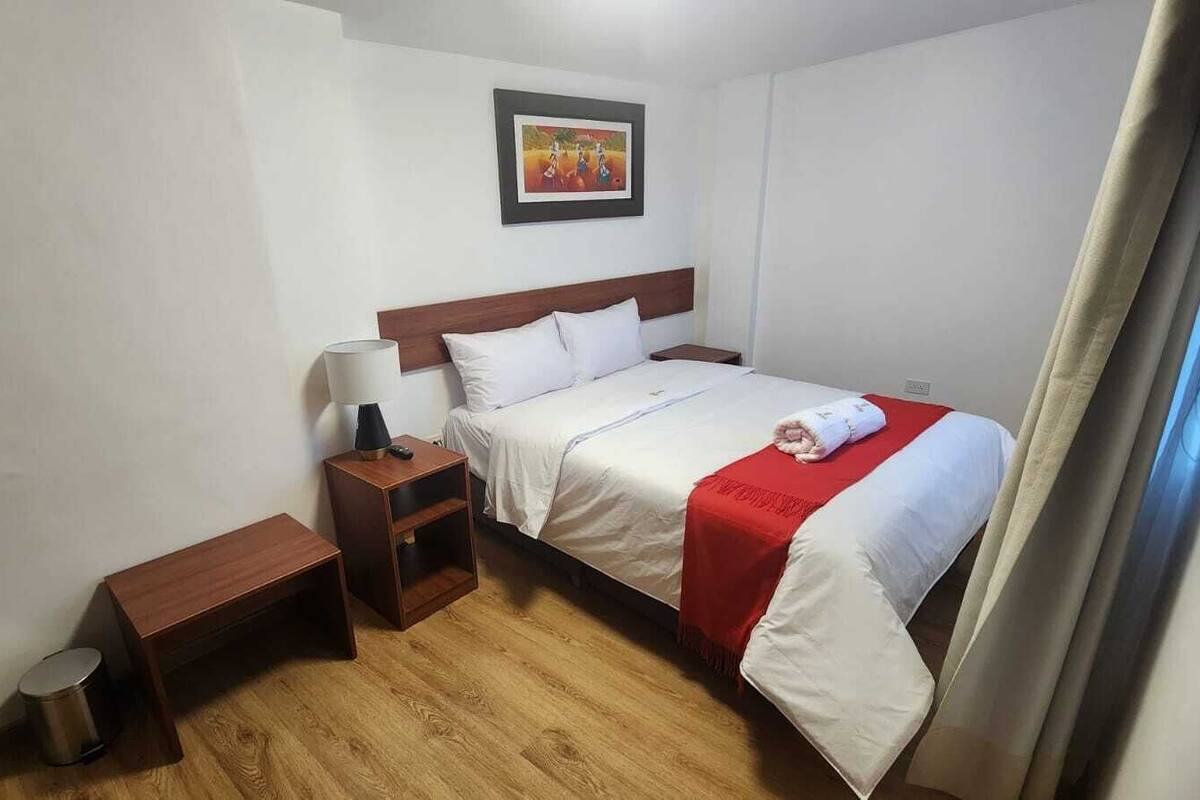A single bed with a wooden headboard, a bedside table with a lamp, a small wooden bench, and a red blanket.