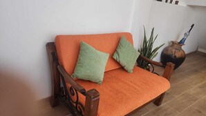 A wooden sofa with orange cushions and green pillows, positioned against a white wall. - INCA PALACE EXPERIENCE (Puno)