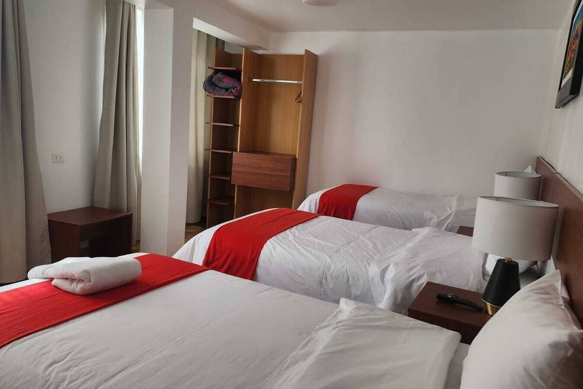 A hotel room with two beds, each with white bedding and red accents, a bedside table with a lamp, and a wardrobe with a mirror.