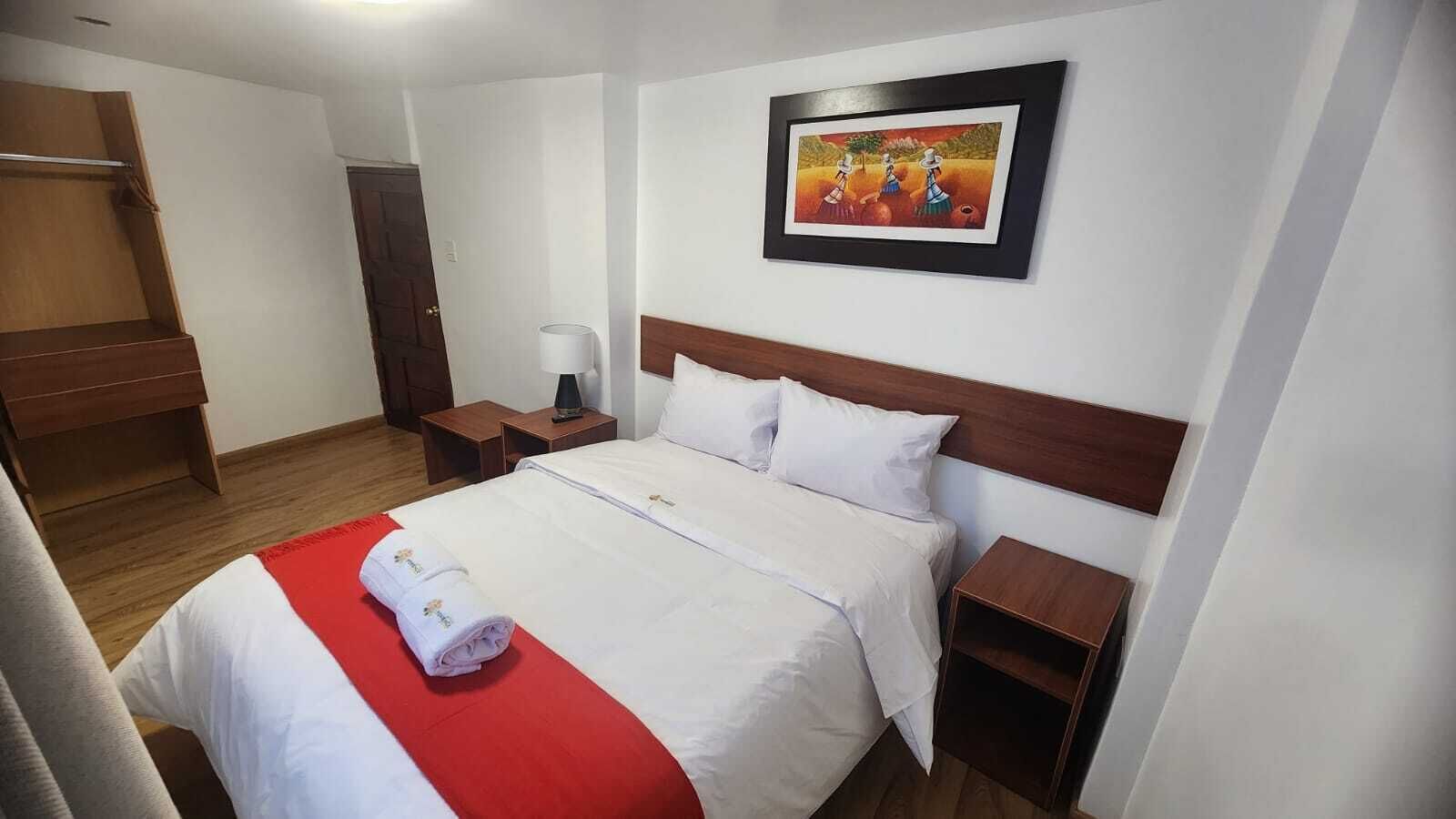 Habitación de hotel con una cama, cabecera de madera, mesita de noche, lámpara y un cuadro en la pared.