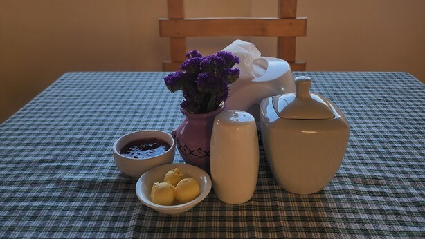 A table with a checkered tablecloth, a vase with purple flowers, a teapot, a salt shaker, a bowl of jam, and a plate with cookies. - INCA PALACE EXPERIENCE (Puno)