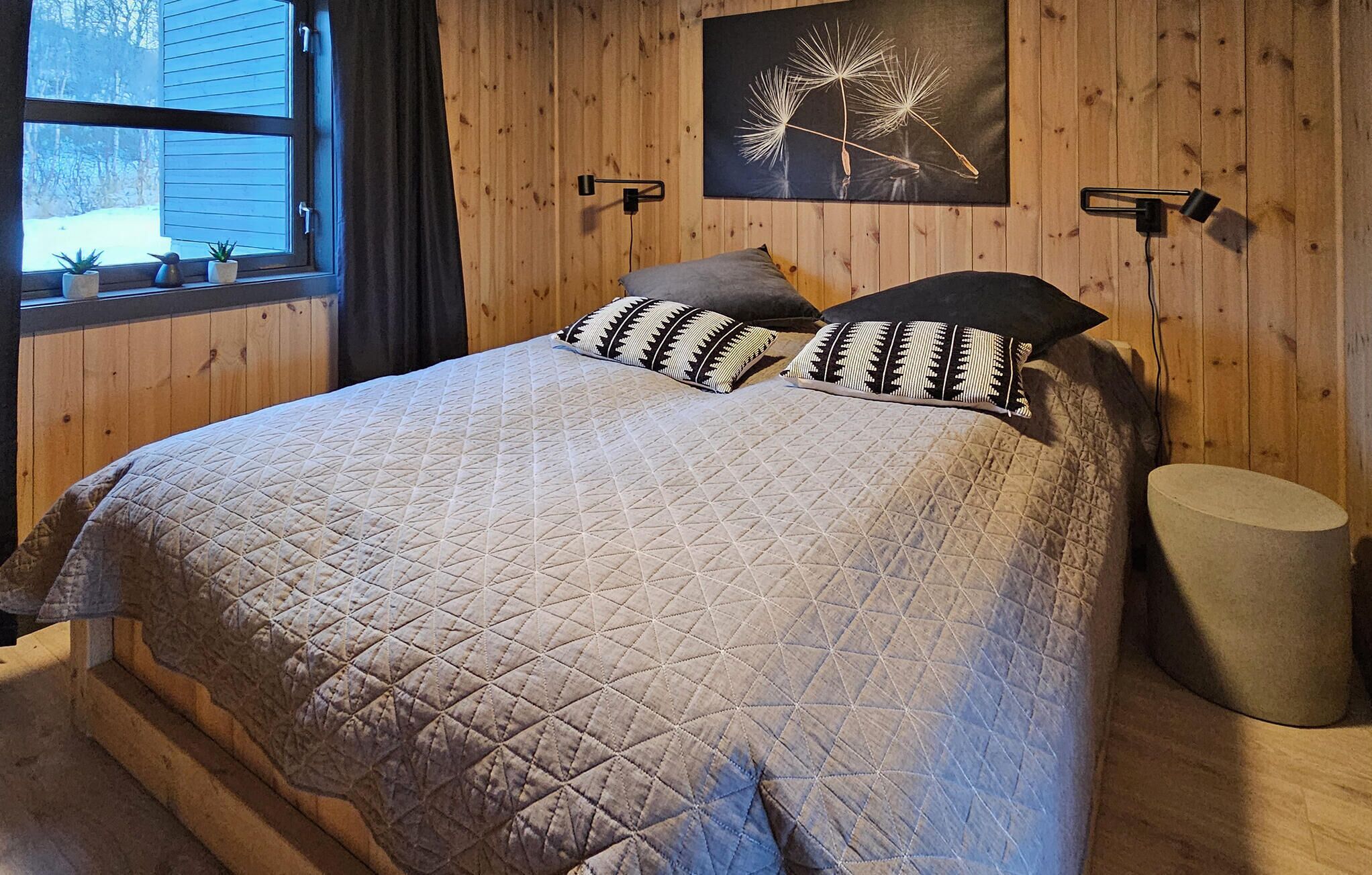 A bedroom with a large bed, two patterned pillows, a wall-mounted lamp, and a window with a view of trees.