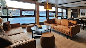 Un salon moderne avec un canapé, un pouf et une table basse, de grandes fenêtres offrant une vue sur un paysage enneigé, et un coin repas avec une table et des chaises.