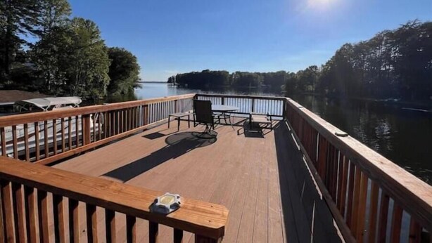 A wooden deck with a table and chairs, overlooking a calm lake with trees in the background.