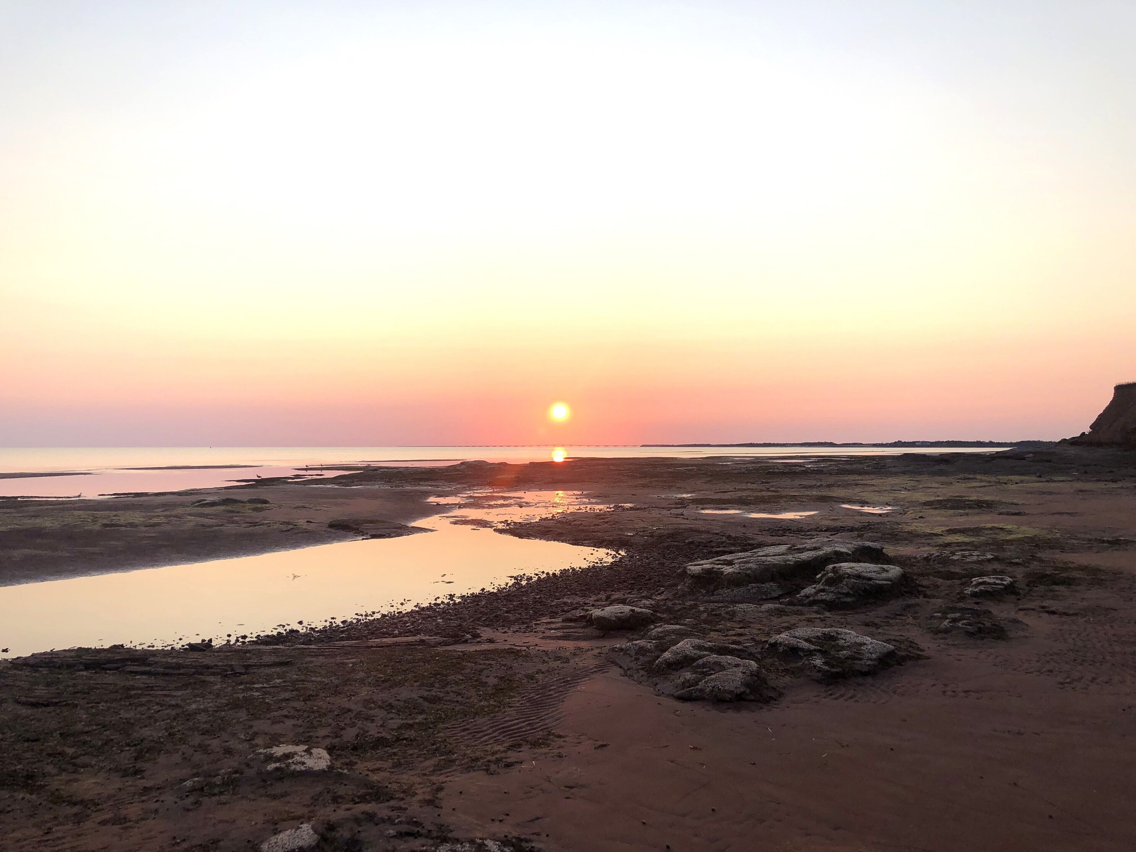 A beach at sunset with a calm sea, rocky terrain, and a distant shoreline.