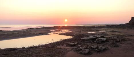 A beach at sunset with a calm sea, rocky terrain, and a distant shoreline.