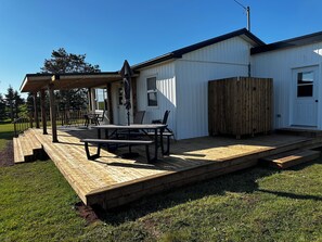Een houten terras met een picknicktafel en stoelen, een overdekt terras en een wit gebouw met een houten hek.