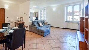 A living room with a sofa, dining table, and chairs. There is a kitchen area in the background with cabinets and a window. - 2 bedroom amazing apartment in Genova (Genova)