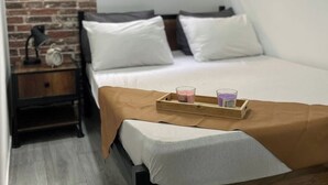 Ein Schlafzimmer mit Backsteinwand, einem Fenster mit Jalousien, einem hölzernen Nachttisch, einem Bett mit weißer Bettwäsche und einem Tablett mit Kerzen.