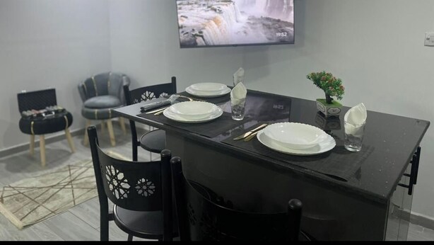 Ein moderner Essbereich mit einem schwarzen Tisch, gedeckt für vier Personen, einem an der Wand befestigten Flachbildfernseher und grauen Stühlen.