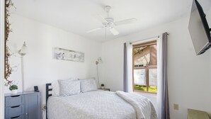 Une chambre à coucher comprenant un lit, des tables de chevet, un ventilateur de plafond, une fenêtre avec des rideaux et une œuvre d’art accrochée au mur.