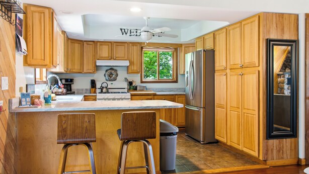 Une cuisine avec des meubles de rangement en bois, un réfrigérateur en acier inoxydable, un tabouret de bar et un ventilateur de plafond.