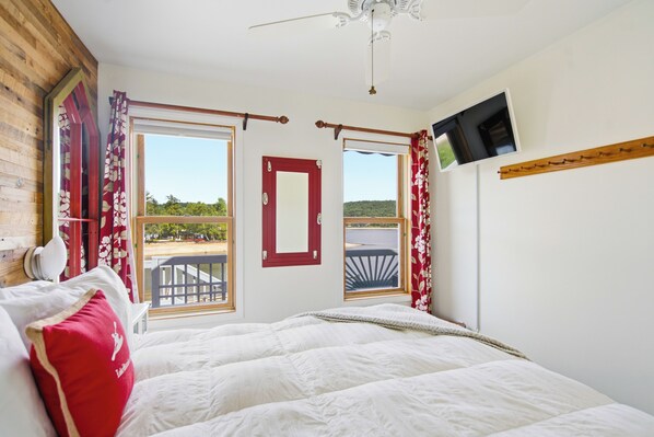 A bedroom with a bed, a TV mounted on the wall, and a view of the outdoors through the windows.