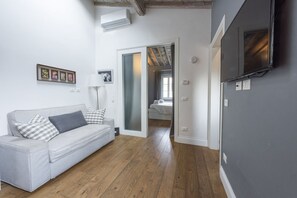 A room with a white sofa, a bed with a white headboard, a TV mounted on the wall, and a wooden floor.