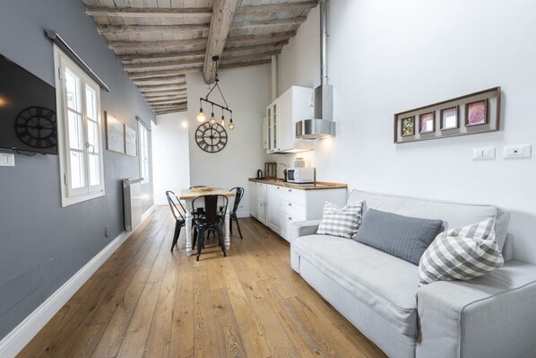 A modern kitchen with wooden flooring, a dining table with chairs, a white sofa with pillows, and a hanging light fixture.