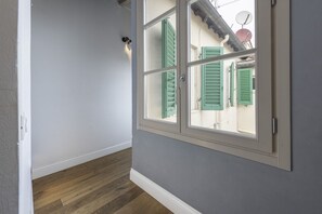 Un couloir avec un plancher en bois, une porte blanche et une fenêtre avec des volets verts.