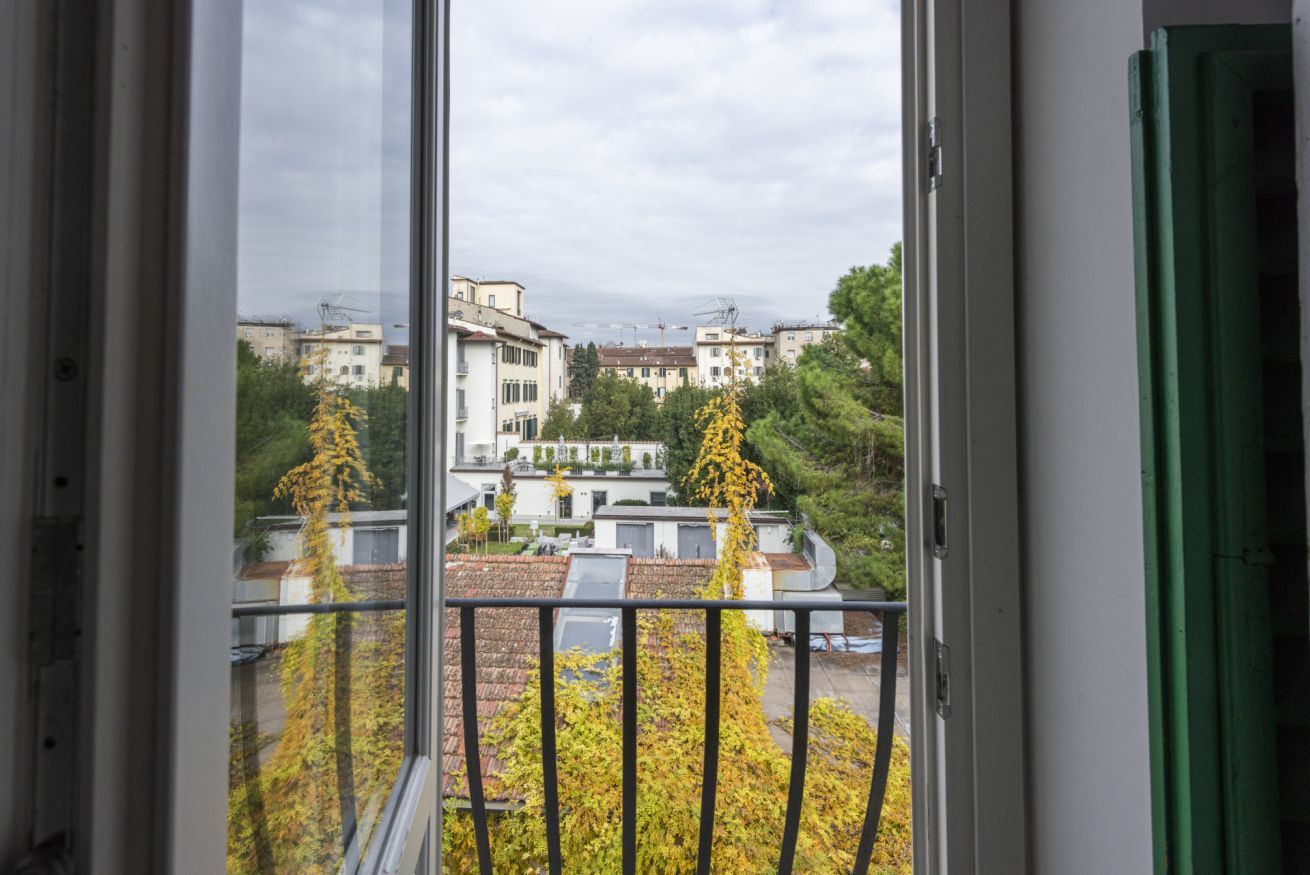 A view from a window with a balcony, featuring residential buildings, trees, and a clear sky.
