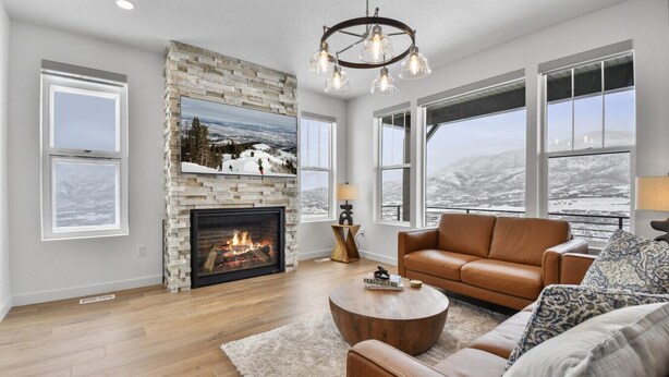 A living room with a fireplace, large windows, a leather sofa, and a chandelier. - Jordanelle Ridge Luxury Retreat (Heber City)