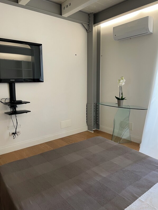 A modern hotel room with a flat-screen TV mounted on the wall, a glass table with a potted orchid, and an air conditioning unit on the wall.