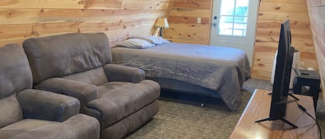 A cozy loft bedroom with a wooden sloped ceiling, a bed, a sofa, a desk with a computer, and a window.