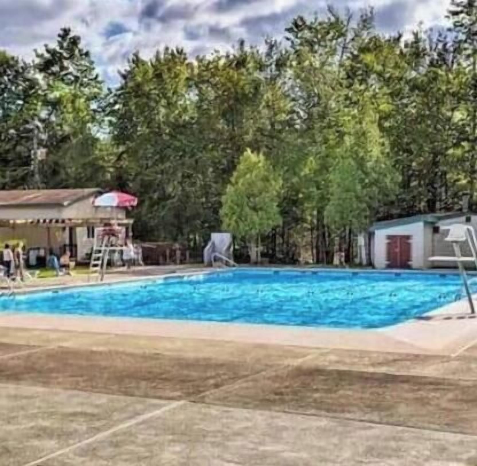 Una piscina con una torre de salvavidas, rodeada de árboles y un edificio con techo rojo.