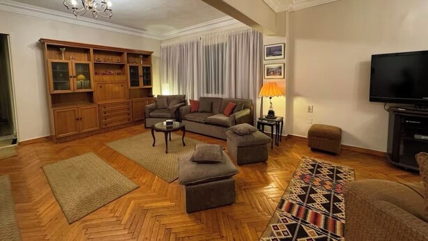Una sala de estar con un conjunto de sofá, un aparador de madera, un televisor y una alfombra estampada.