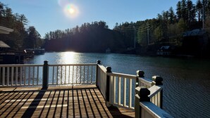 A wooden deck with a railing overlooking a calm lake, surrounded by trees and buildings. - Green Acres – Your lakefront place to be! (Lakemont)