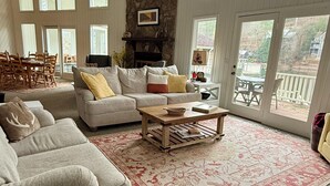 A spacious living room with a stone fireplace, large windows, and a cozy seating area. - Green Acres – Your lakefront place to be! (Lakemont)