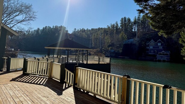 A wooden deck with a gazebo, overlooking a lake with a house and trees in the background. - Green Acres – Your lakefront place to be! (Lakemont)