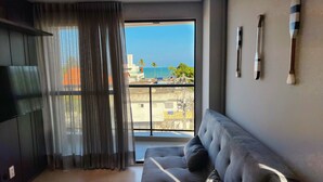 A modern living room with a large window, a grey sofa, and three paddleboard-like decorations on the wall. - Oceânica Cabo Branco (João Pessoa)