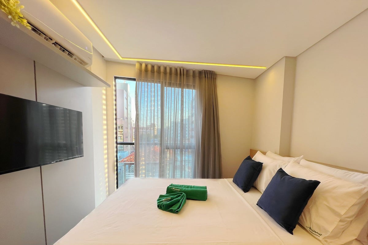 A hotel room with a bed, a flat-screen TV, a window with curtains, and a view of buildings outside.