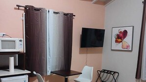 Een kleine, goed verlichte kamer met een bed, een bureau met een magnetron, twee stoelen, een televisie en een schilderij aan de muur.